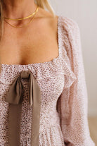 Close-up of a woman wearing a light pink and white micro-print top with a square neckline, ruffled trim, and a large taupe bow at the center front. She is also wearing a layered gold chain necklace.