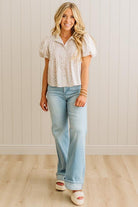Woman wearing a white patterned blouse and light blue jeans standing against a white paneled wall.