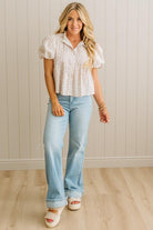 Woman wearing a white patterned blouse and light blue jeans against a neutral wall.