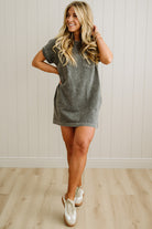 Woman wearing a gray t-shirt dress with white sneakers against a light wooden floor and wall background.