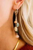 A close-up of a blonde woman wearing a dangling statement earring with irregular polished stones in peach, green, cream, and dark green, suspended from gold-toned hardware.