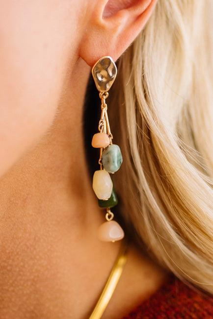 A close-up of a blonde woman wearing a dangling statement earring with irregular polished stones in peach, green, cream, and dark green, suspended from gold-toned hardware.