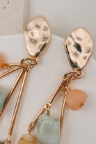 Close-up of a pair of gold-toned dangle earrings with hammered teardrop-shaped studs, thin dangling bars, and small peach and pale green stone charms on a soft white background.