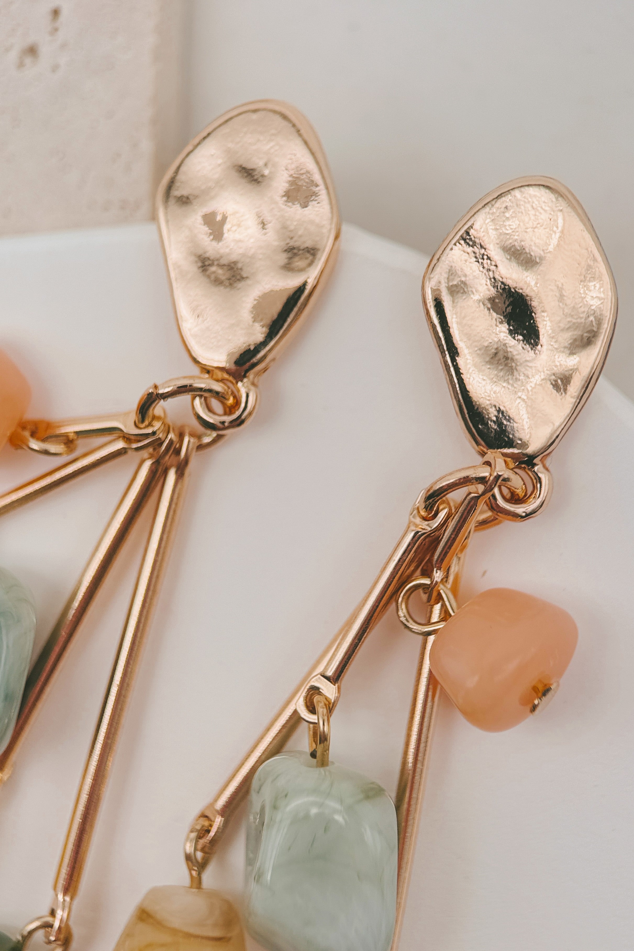 Close-up of a pair of gold-toned dangle earrings with hammered teardrop-shaped studs, thin dangling bars, and small peach and pale green stone charms on a soft white background.