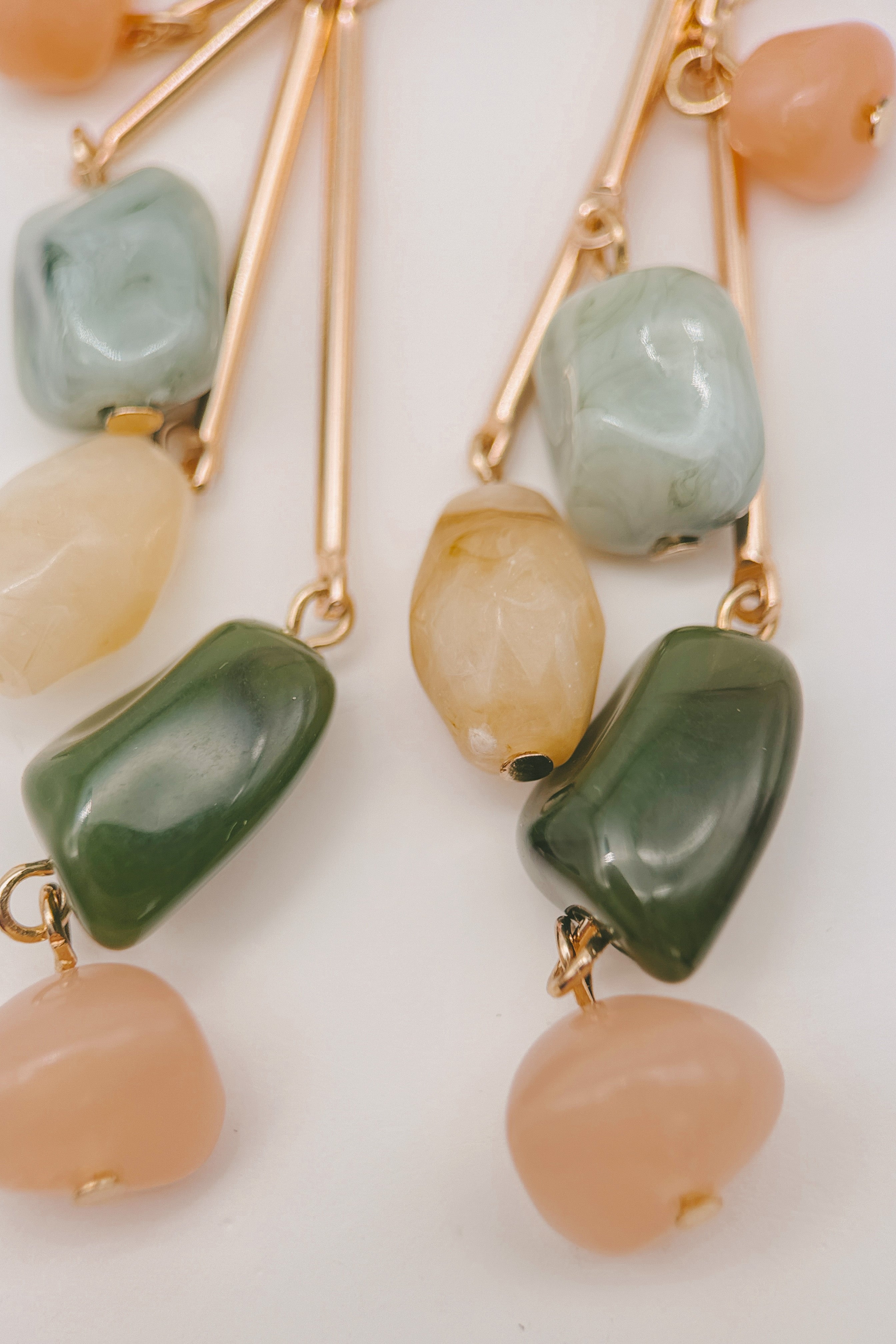 Close-up of dangling earrings with gold-tone metal and irregular polished beads in pale green, dark green, cream, and peach on a white background.