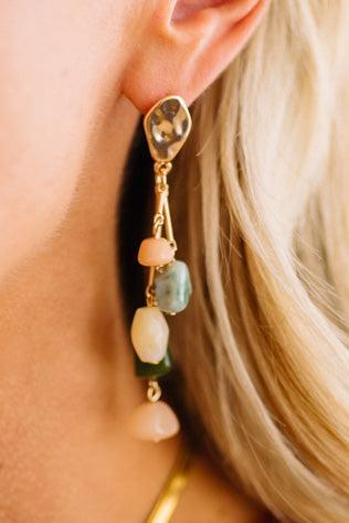 A close-up of a model's ear wearing a dangling gold earring with irregular pastel gemstone beads in pink, green, and cream.