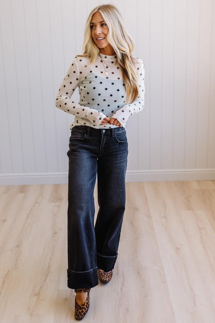 Woman wearing a polka dot top and jeans standing in a room with wooden flooring and white walls.