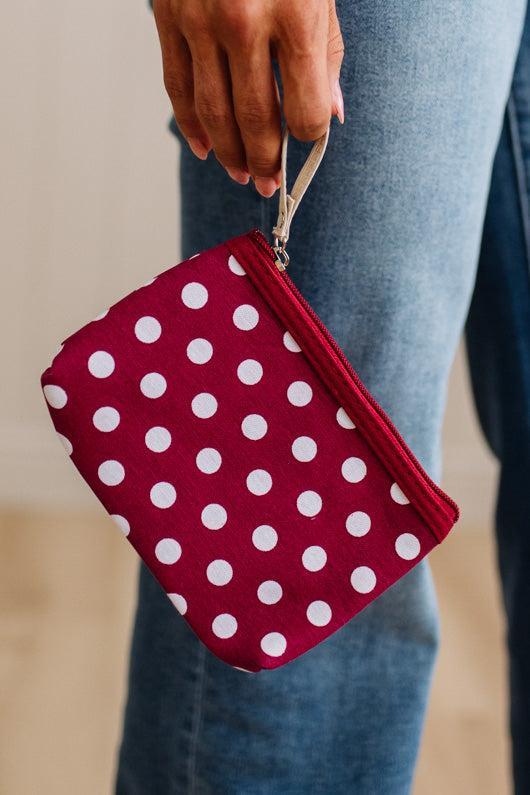 A close-up of a person holding a small zip pouch with a wrist strap. The pouch is burgundy red with white polka dots and has a silver metal clip.