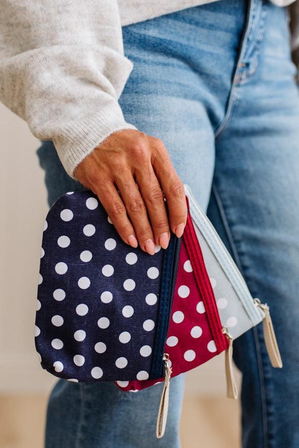 Three small zip pouches with white polka dots are arranged vertically on a light wood surface. The top pouch is pale blue, the middle is navy blue, and the bottom is red, each with a small beige zipper pull.