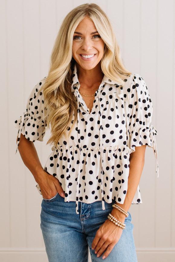 A woman wearing a white short-sleeve blouse with black polka dots, tied ruffle cuffs, and a peplum waist, paired with blue jeans, standing against a white paneled wall.
