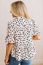 A woman viewed from the back wearing a white short-sleeve blouse with black polka dots, ruffled sleeves, and a gathered peplum hem, paired with blue jeans against a white paneled wall.