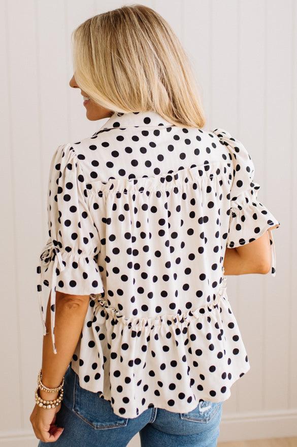 A woman viewed from the back wearing a white short-sleeve blouse with black polka dots, ruffled sleeves, and a gathered peplum hem, paired with blue jeans against a white paneled wall.