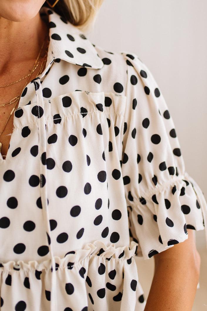 Close-up of a woman wearing a cream-colored blouse with black polka dots, featuring a pointed collar, gathered chest details, and ruffled short sleeves.