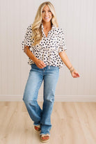 A woman modeling a white short-sleeve blouse with black polka dots, paired with light-wash blue jeans and tan sandals, standing against a white paneled wall and light wood floor.