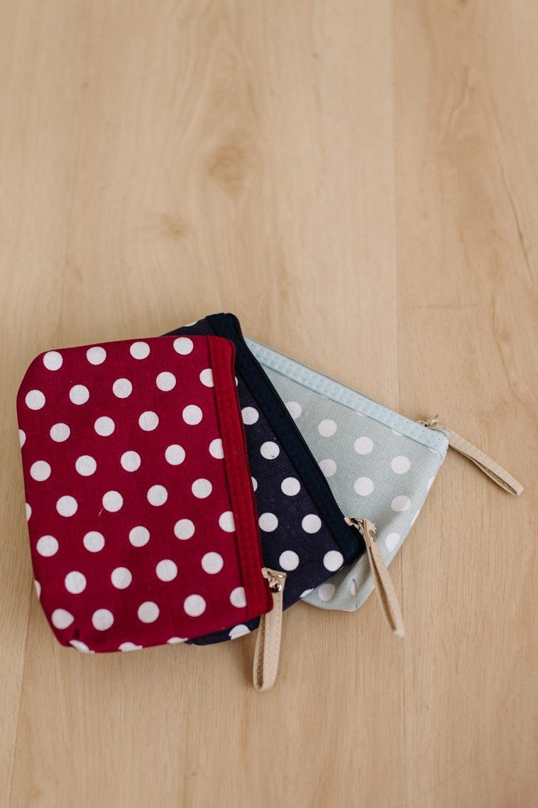 Three small zippered pouches with white polka dots are arranged on a light wood surface. The front pouch is red with white polka dots, partially covering a navy blue pouch and a pale blue pouch.