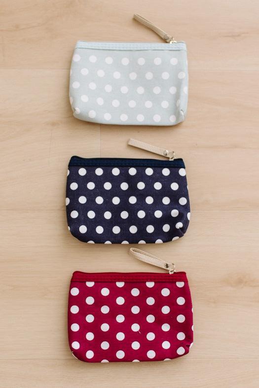 Three small zip pouches with white polka dots are arranged vertically on a light wood surface. The top pouch is pale blue, the middle is navy blue, and the bottom is red, each with a small beige zipper pull.