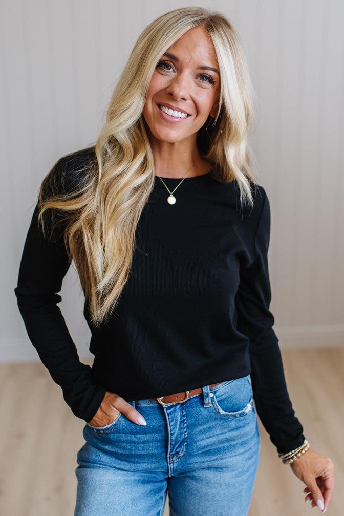 A smiling blonde woman wearing a black long-sleeve crewneck top and blue jeans, standing in a brightly lit indoor setting with light-colored walls and wood flooring.