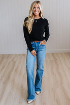 A woman stands in a bright room with light wood floors and white paneled walls, wearing a black long-sleeve top, blue wide-leg jeans, and beige pointed shoes.