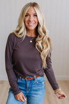 A smiling blonde woman wearing a dark brown long-sleeve crewneck top, blue jeans, and a brown belt, standing against a light beige paneled wall.