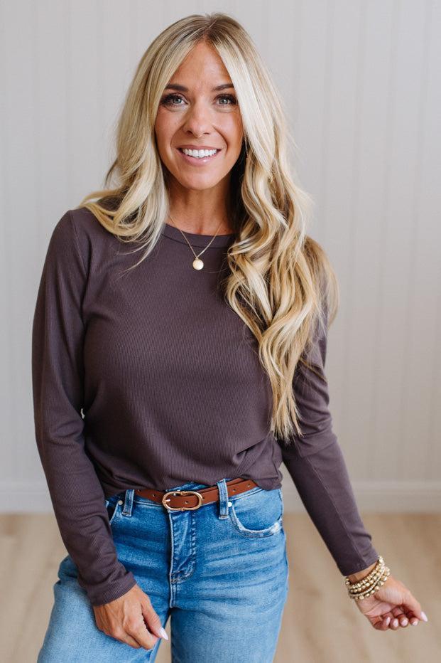 A smiling blonde woman wearing a dark brown long-sleeve crewneck top, blue jeans, and a brown belt, standing against a light beige paneled wall.