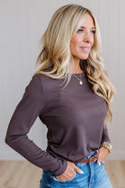 A woman with blonde hair wearing a long-sleeve crewneck top in a muted plum-brown color, paired with blue jeans and a brown belt, posing in a brightly lit indoor setting.