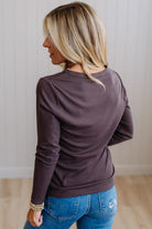 A woman with blonde hair is shown from the back wearing a dark brown long-sleeve crewneck top and blue jeans, standing in a room with light-colored paneled walls and a wood floor.