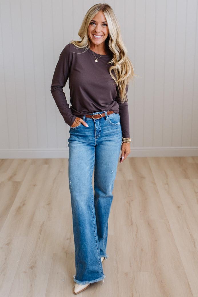 A woman modeling a dark brown long-sleeve knit top and blue flare jeans with a brown leather belt, standing in a room with light wood floors and white paneled walls.