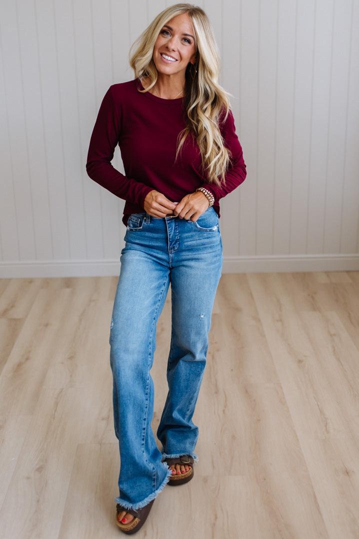 A woman modeling a long-sleeve burgundy top and blue jeans, standing on a light wood floor against a white paneled wall.