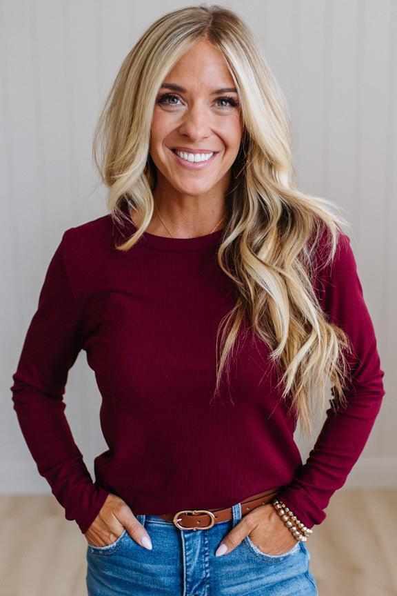 A smiling blonde woman wearing a long-sleeve crewneck top in deep burgundy, paired with blue jeans and a brown belt, standing against a light neutral wall.
