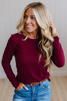 A smiling blonde woman wearing a fitted long-sleeve crewneck top in deep burgundy, paired with blue jeans and a brown belt, standing against a light neutral wall.