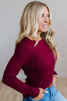 A woman with blonde hair wearing a long-sleeve crewneck top in deep burgundy, paired with blue jeans, posing in a brightly lit indoor setting.