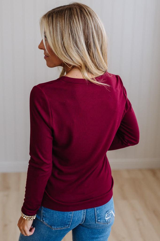 A woman with blonde hair is shown from the back wearing a long-sleeve crewneck top in deep burgundy, paired with blue jeans, in an indoor setting with light wood floors and a white paneled wall.