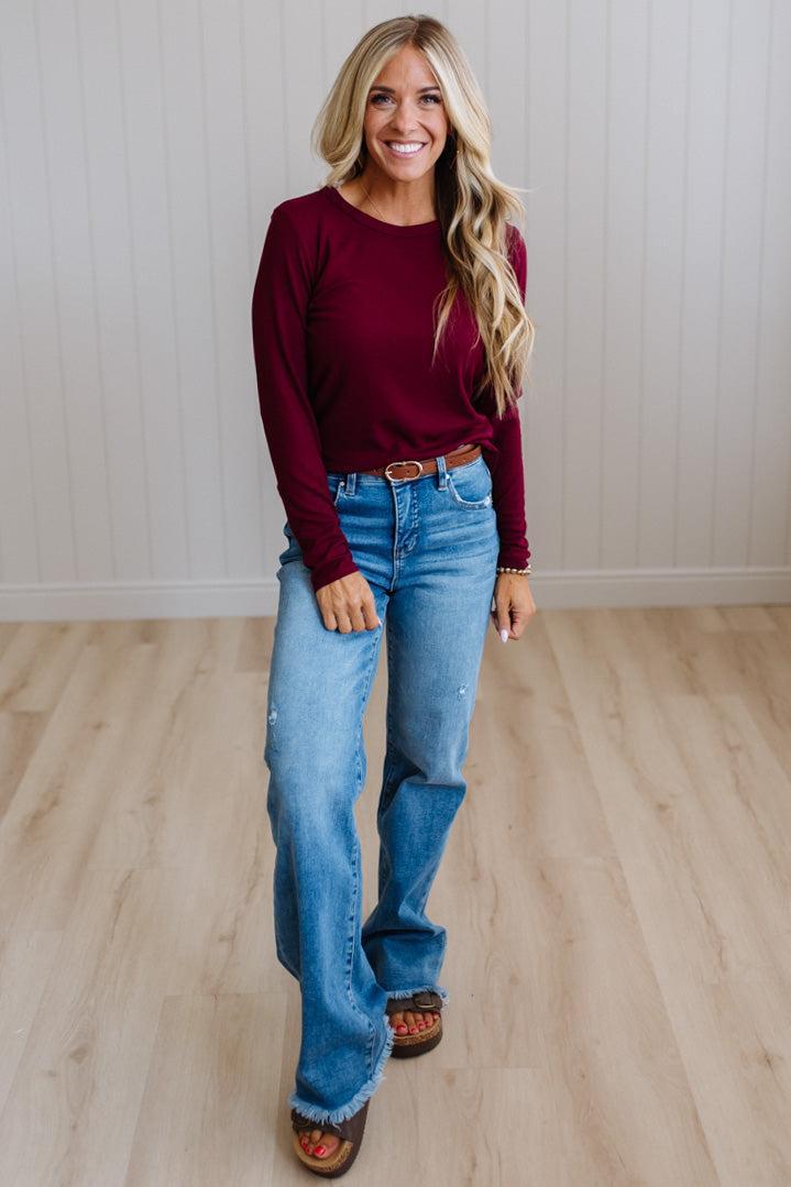 A woman modeling a long-sleeve crewneck top in burgundy, paired with blue jeans and brown sandals, standing in a room with light wood floors and white paneled walls.