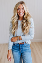 A smiling blonde woman wearing a light gray long-sleeve crewneck top, blue jeans, and a brown belt, standing in a bright room with light wood floors and white paneled walls.