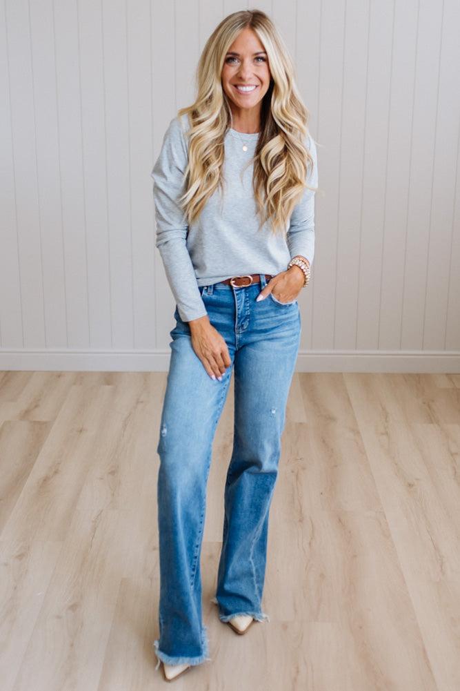 A woman modeling a light gray long-sleeve knit top and blue flare jeans with a brown leather belt, standing in a room with light wood floors and white paneled walls.