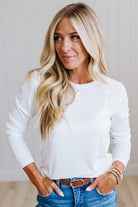 A woman with long blonde hair wearing a white long-sleeve crewneck top and blue jeans, standing against a light-colored paneled wall.