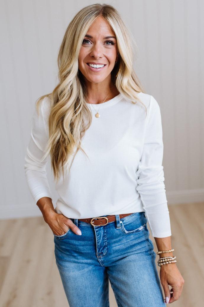 A smiling blonde woman wearing a white long-sleeve crewneck top, blue jeans, and a brown belt, standing in a brightly lit room with light wood floors and white paneled walls.