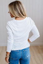 A woman viewed from behind wearing a white long-sleeve crewneck top and blue jeans, standing in a brightly lit room with light wood flooring and white paneled walls.