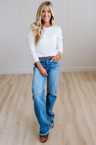 A woman modeling a white long-sleeve henley shirt and wide-leg blue jeans with a brown belt, standing on a light wood floor against a white paneled wall.