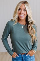 A smiling blonde woman wearing a muted sage green long-sleeve top and blue jeans, standing with her hands in her pockets against a light neutral wall.