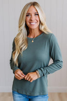 A smiling blonde woman wearing a muted sage green long-sleeve top and blue jeans, standing against a light paneled wall.