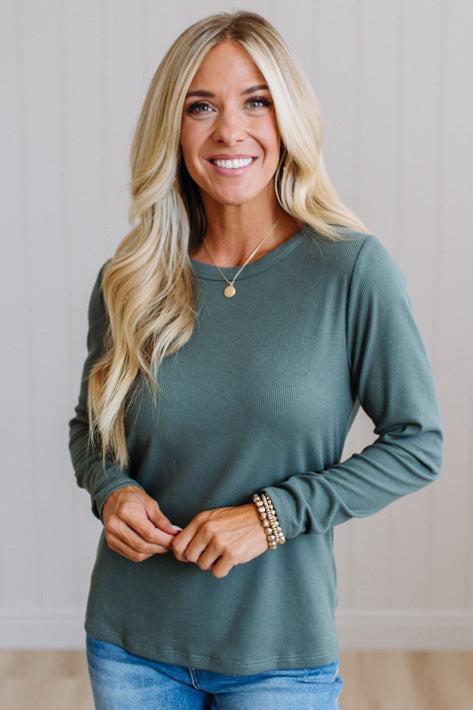 A smiling blonde woman wearing a muted sage green long-sleeve top and blue jeans, standing against a light paneled wall.