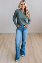 A woman modeling a sage green long-sleeve crewneck top and blue wide-leg jeans, standing in a room with light wood floors and white paneled walls.