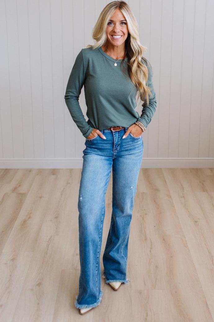 A woman modeling a sage green long-sleeve crewneck top and blue wide-leg jeans, standing in a room with light wood floors and white paneled walls.