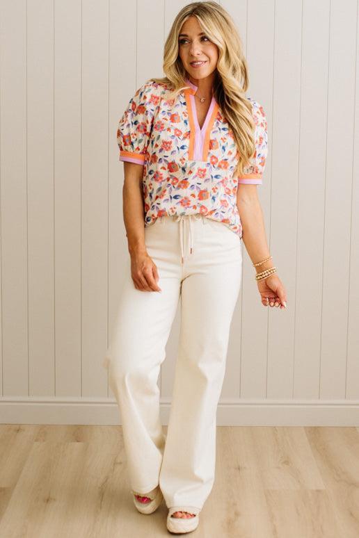 Woman wearing a floral blouse and white pants against a light wooden paneled wall.