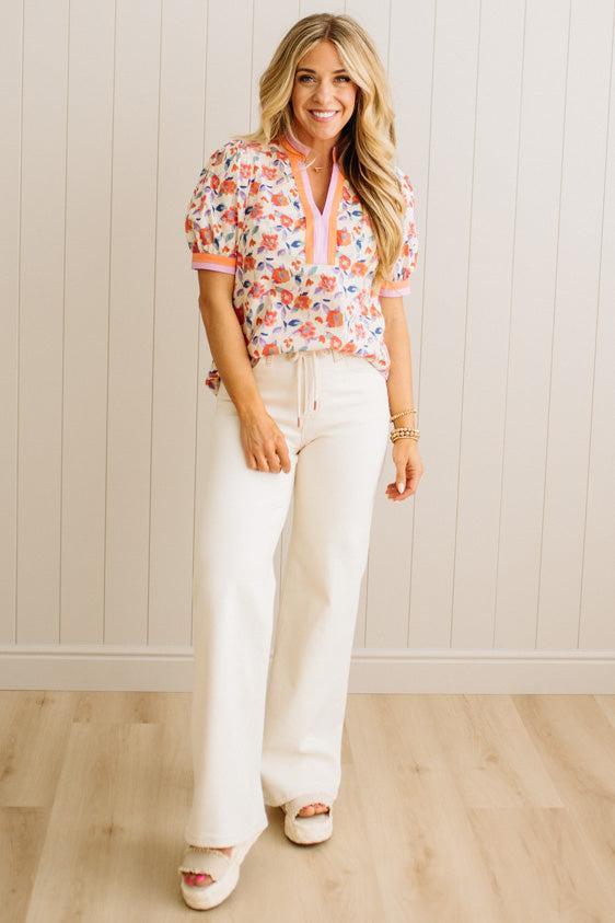 Woman wearing a floral blouse and white pants against a light wooden paneled wall.