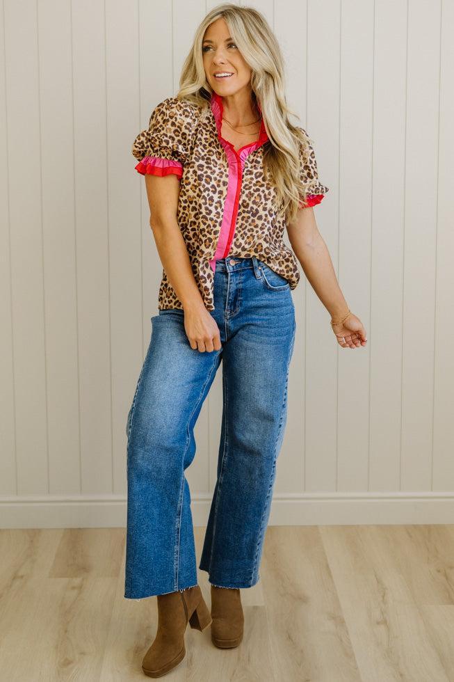Woman wearing a leopard print shirt with red trim and blue jeans against a white paneled wall.