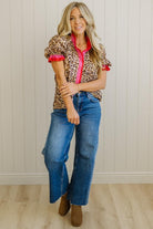 Woman wearing a leopard print top with red trim, blue jeans, and brown boots in a room with a wooden floor and white paneled walls.