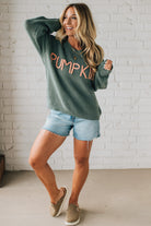 Woman wearing a green 'Pumpkin' sweater with denim shorts against a white brick wall.