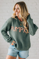 Woman wearing a green sweater with 'PUMPKIN' text, standing against a white brick wall.